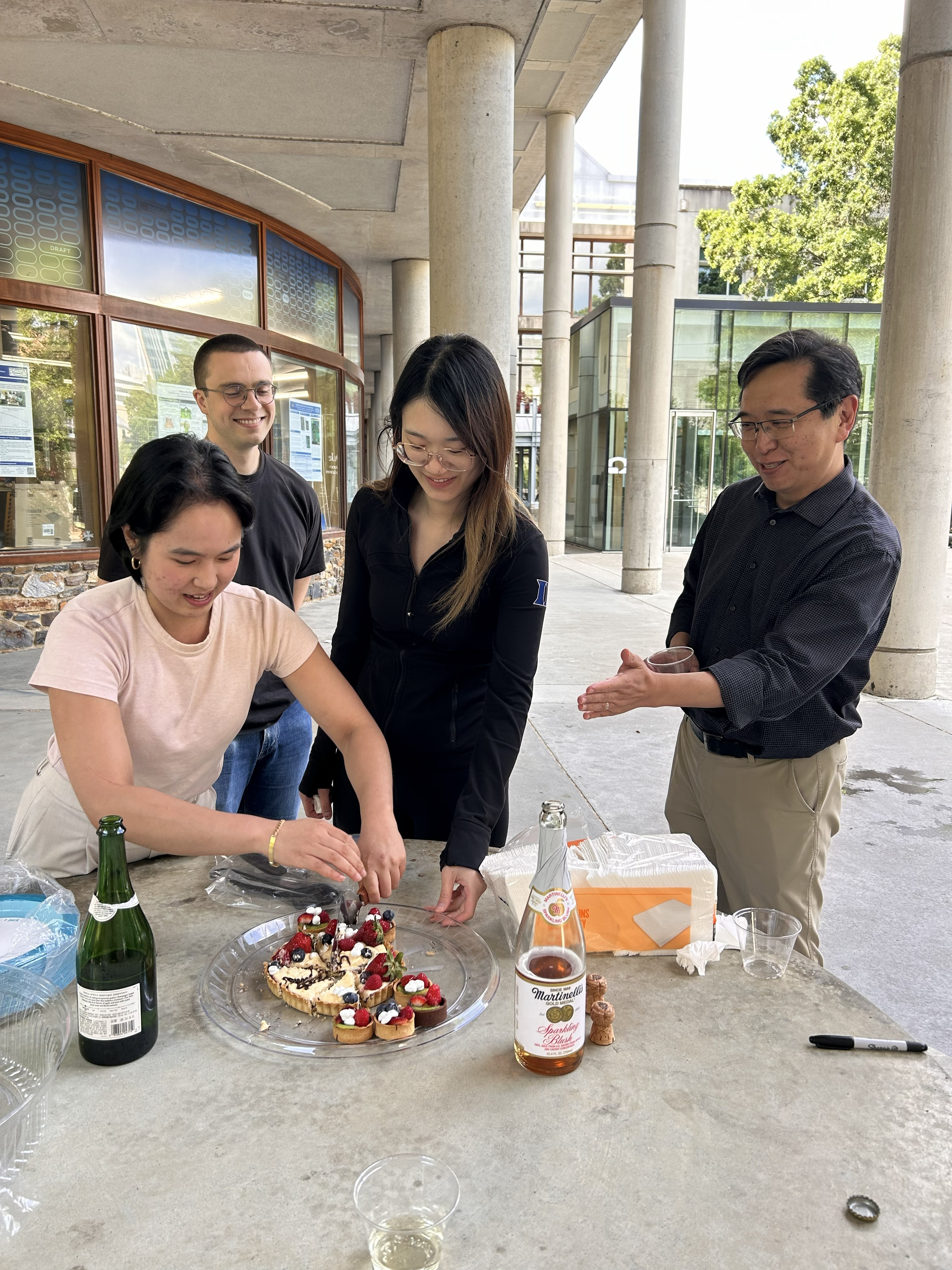 Mariel De Los Santos cuts tart with Nolan Bick, Xing Gao, and Lee Zou observing in celebration of successful preliminary exams.