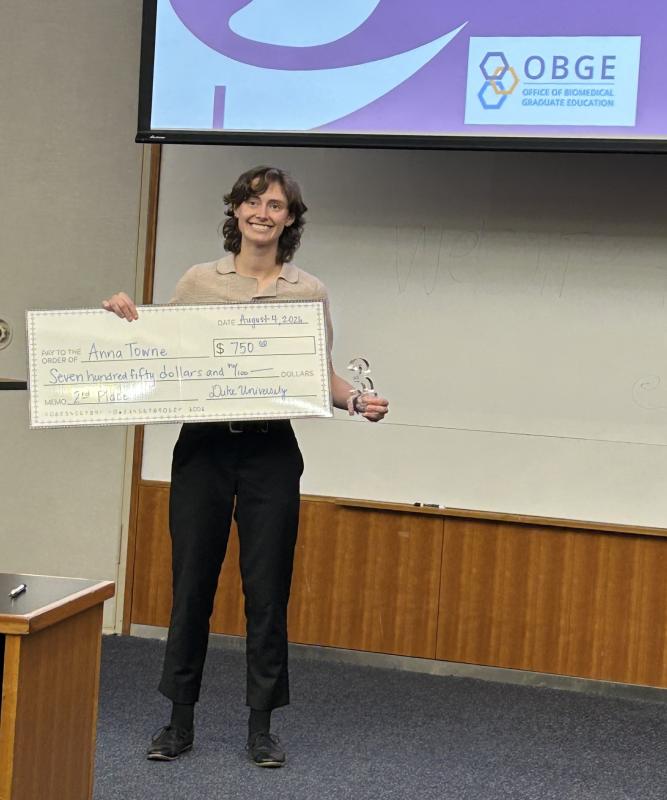 Anna Towne stands at the front of a lecture hall holding an oversized ceremonial check for $750 and a clear, number-shaped trophy displaying a “3” for her 3MT award. A presentation screen above shows the Office of Biomedical Graduate Education (OBGE) logo. The check is dated August 4, 2026, and awarded by Duke University.