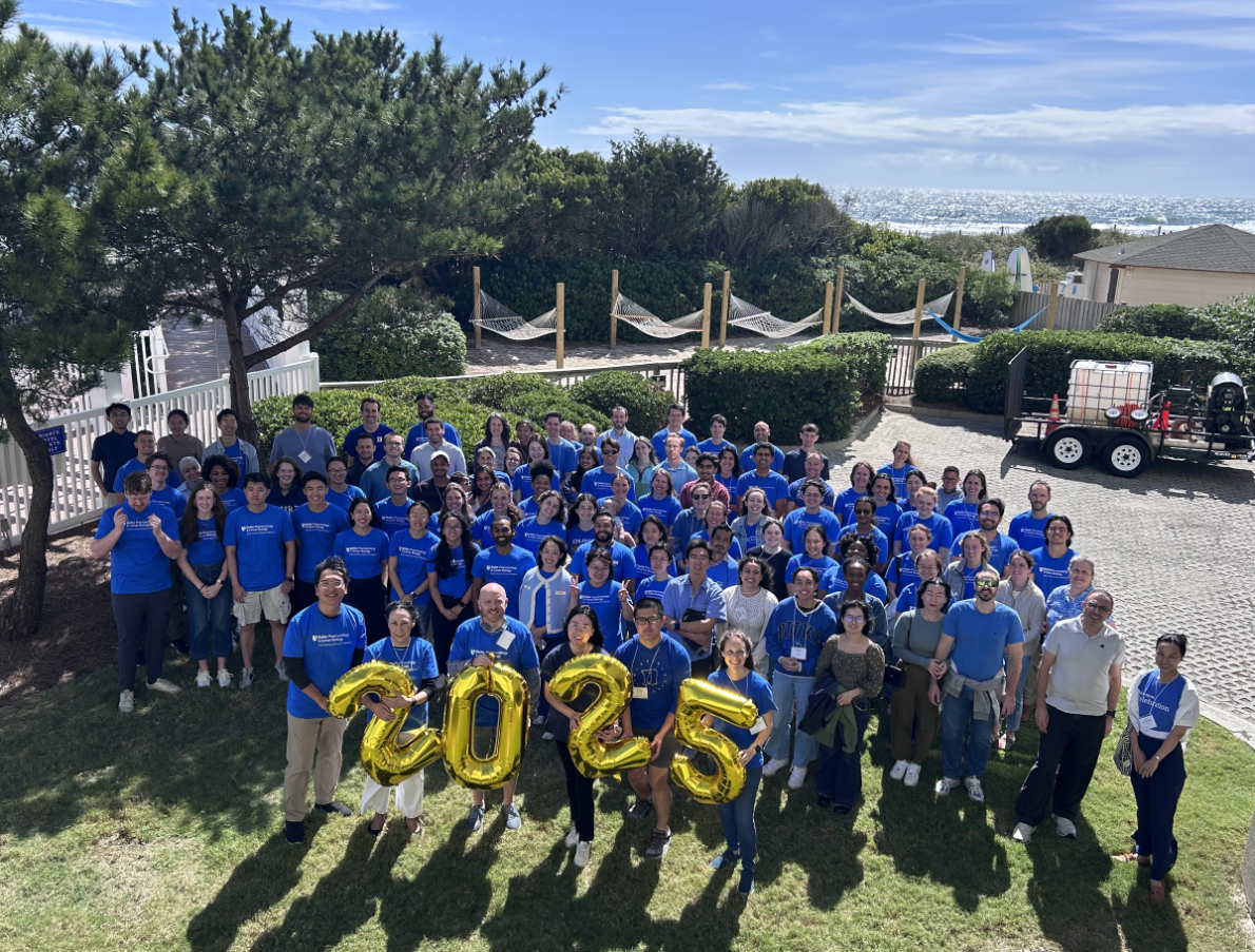 Group photo of PCB department retreat attendees standing outside on a sunny day at Wrightsville Beach. Four attendees hold gold balloons spelling out 2025.